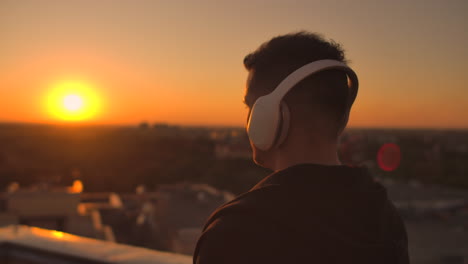 A-man-walks-on-the-roof-at-sunset-with-headphones-looking-at-the-city-from-the-height-of-a-skyscraper-at-sunset.-Relax-while-listening-to-music.-Enjoy-a-beautiful-view-of-the-city-at-sunset-from-the-roof-with-headphones